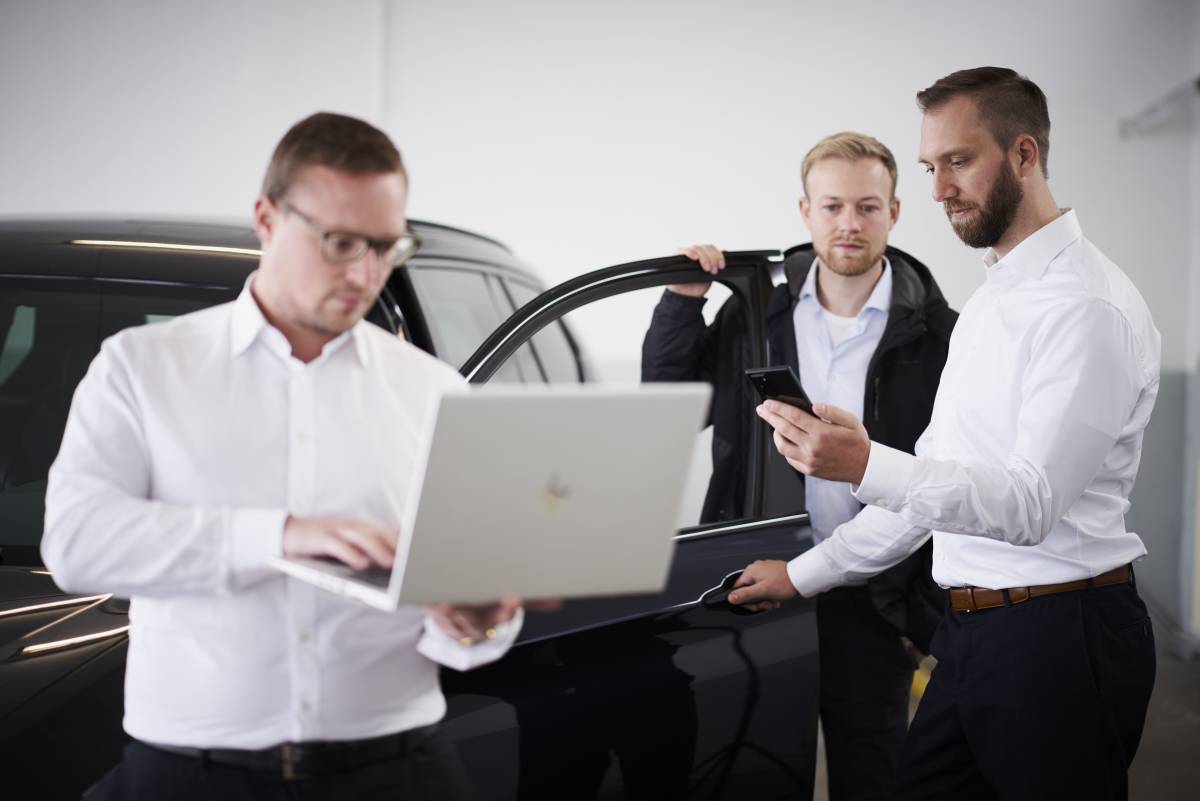 Three people standing around a car with an open door, holding a laptop and a smartphone